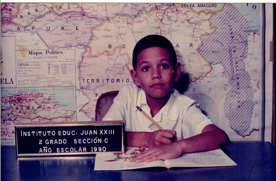A boy of seven at a school desk, pencil in hand over an open exercise book, in front of a wall-sized political map of Venezuela. A letterboard on the desk reads Instituto Educ. Juan XXIII, 2 grado sección C, año escolar 1990.