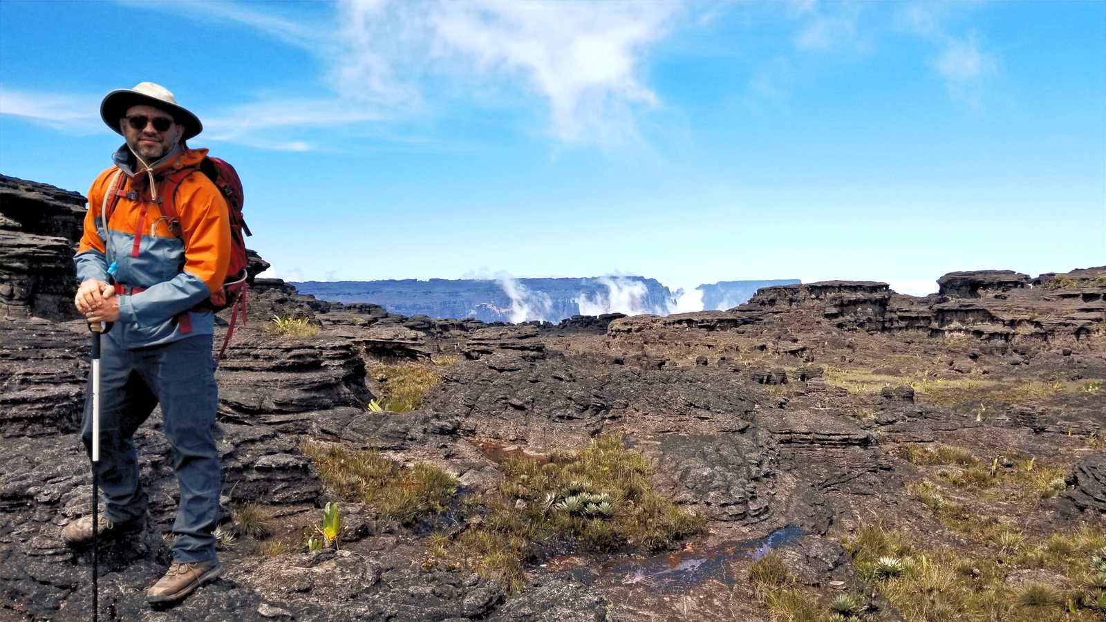 Standing on the bare rock summit plateau of a tepui, walking pole in hand, with cloud spilling over the escarpment behind.
